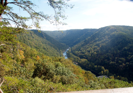 [The river cuts through the valley coming around the bend to a straighter section with has a bridge just above the water level. The hillsides are completely filled with trees except for a section on the right side of the river which is open for the road alongside the river. This view is high enough that not all of the river is visible and just the top of the bridge can be seen.]
